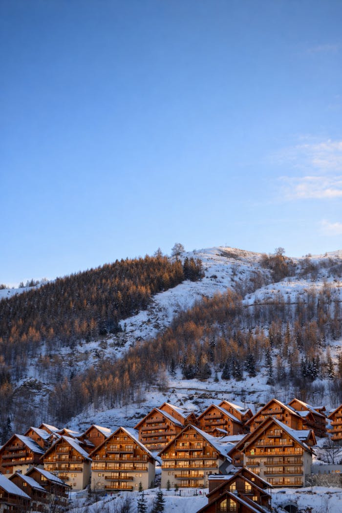 Chalets de montagne enneigés au pied d’une colline boisée, baignés par la lumière douce de la fin de journée sous un ciel bleu d’hiver